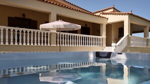 Une piscine dont l’eau est si claire qu’elle reflète l’environnement, un balcon couvert agrémenté d’une balustrade blanche et un bâtiment surmonté d’un toit en tuiles.