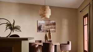 Un coin repas comprenant une table ronde, six chaises, un luminaire suspendu en fibres tressées, un grand tableau abstrait et une plante en pot.