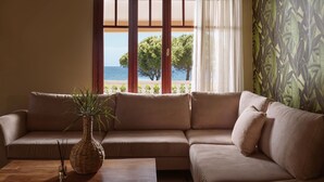 Un salon avec un canapé beige, une table basse en bois, un vase en fibres naturelles contenant une plante, et une vue sur la mer grâce à de grandes fenêtres ornées de rideaux.