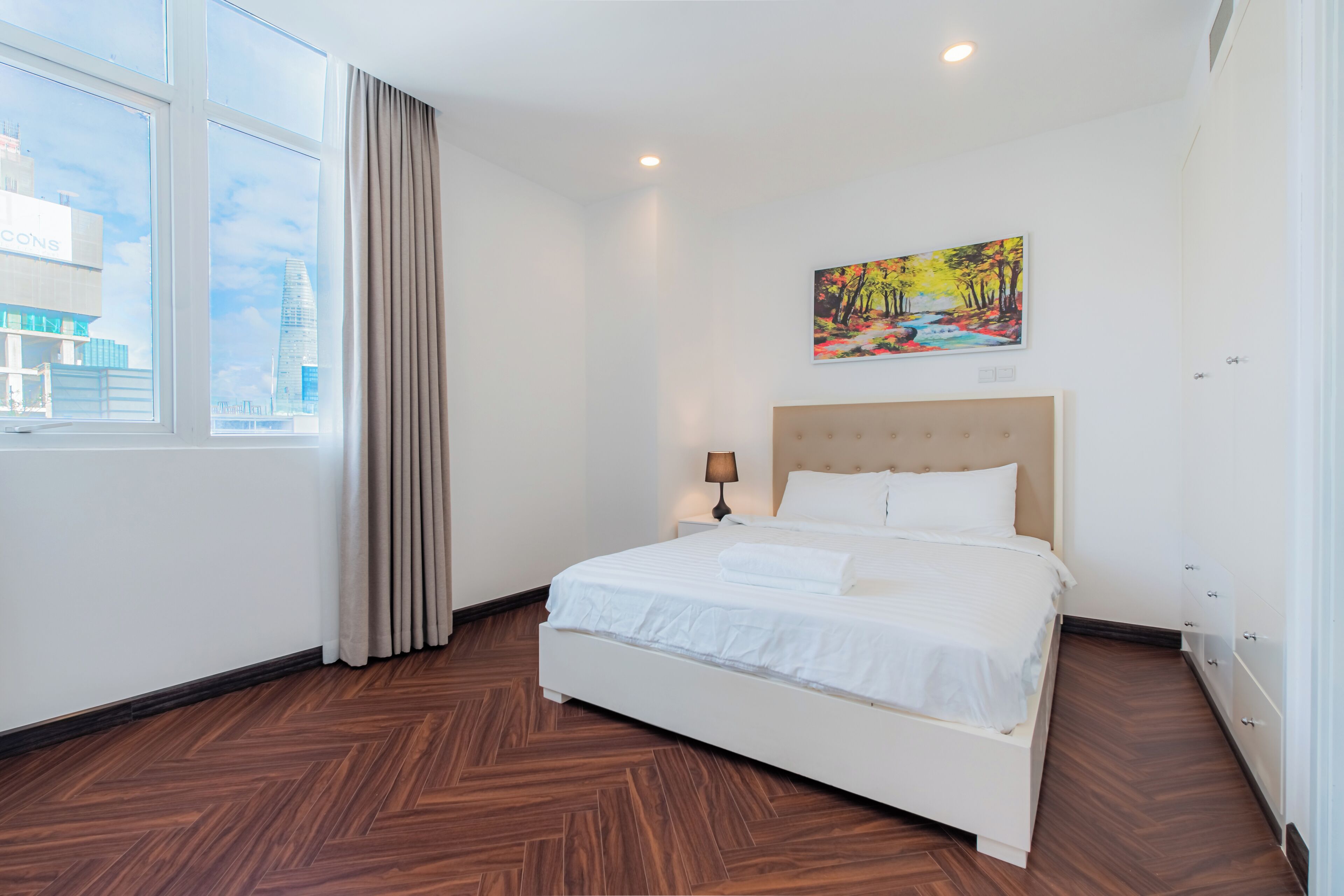 A hotel room with a large bed, a painting on the wall, a window with curtains, and a view of buildings outside.