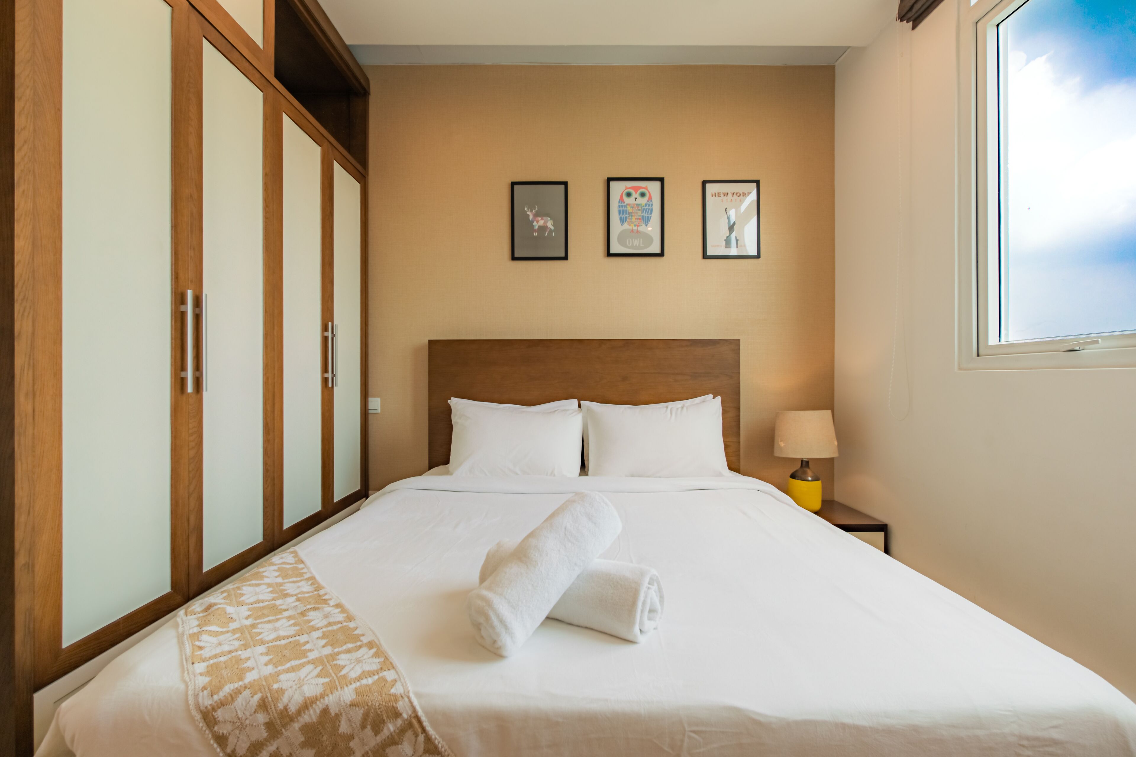 A neatly made bed with white linens and a rolled towel, a wooden wardrobe, and a nightstand with a lamp.