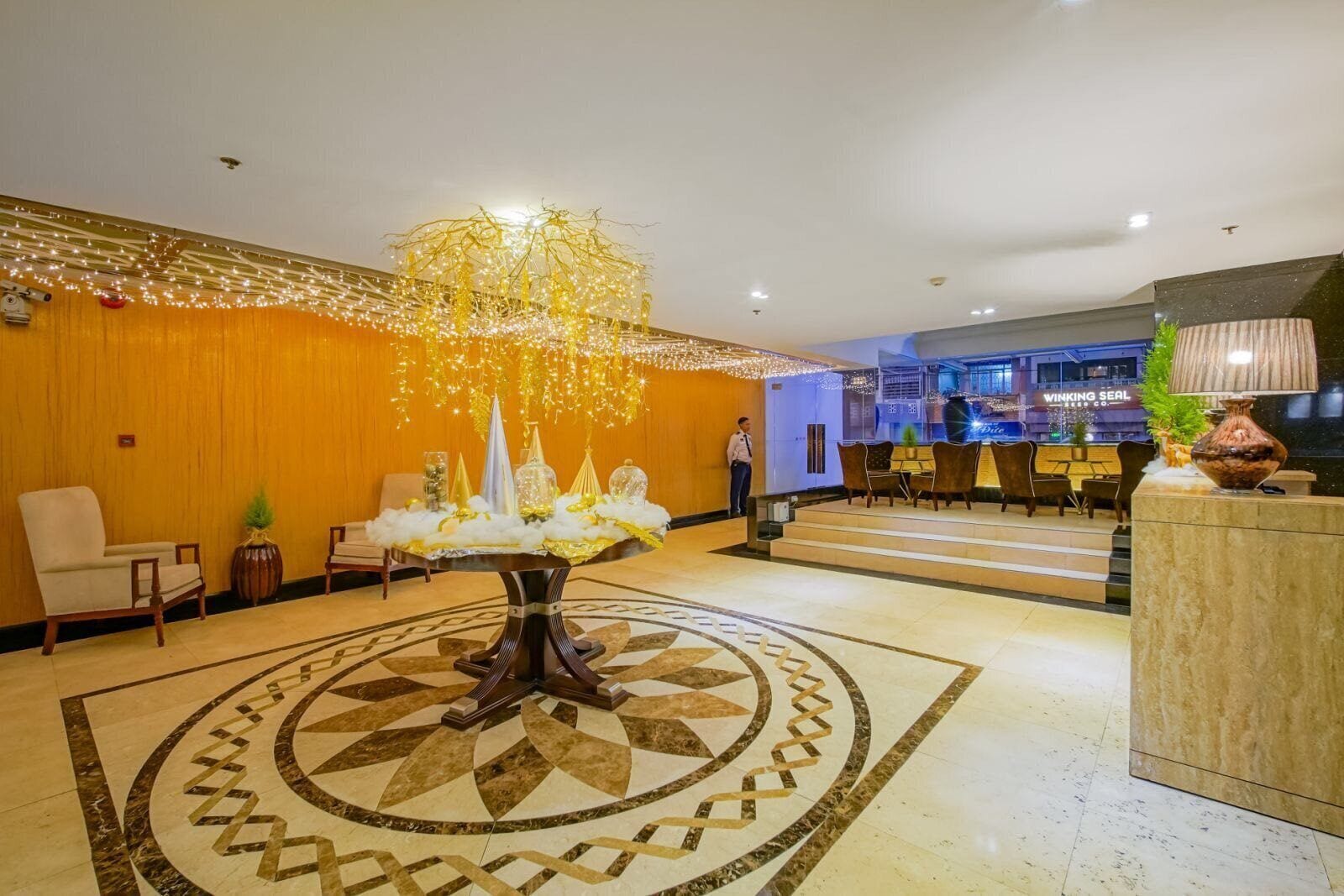 Un vestíbulo con una fuente decorativa central, un mostrador de recepción y un área de descanso con sillas y mesas.