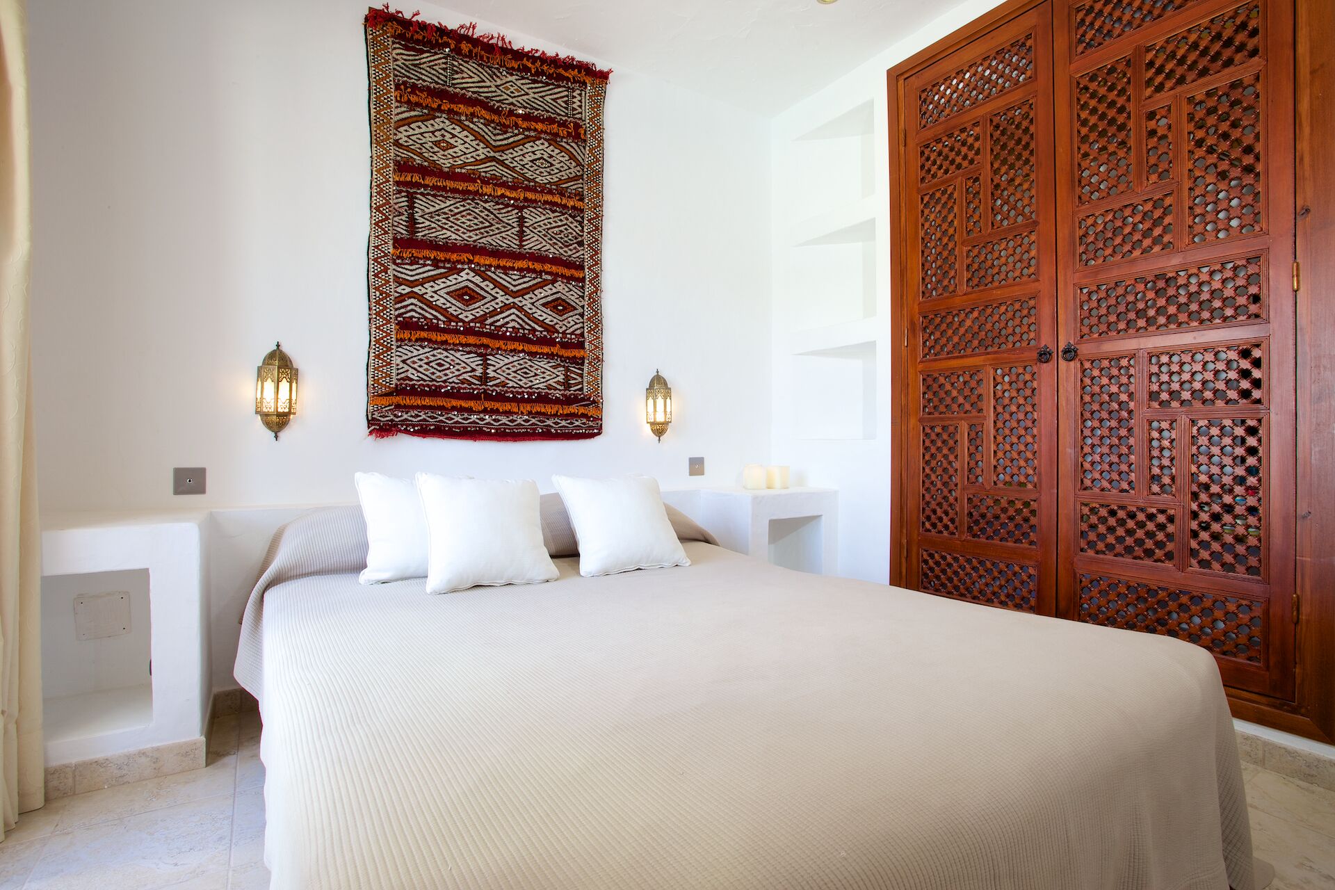 A bedroom with a bed, two bedside tables, a wall-mounted tapestry, and a wooden door with intricate lattice design.