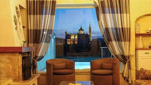 A room with two brown armchairs, a small table with a plate of food, a fireplace, and a view of a historic building through a window with striped curtains. - Twilight Holiday Home (Għasri)