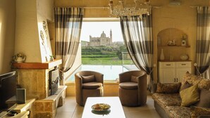A living room with a stone fireplace, two armchairs, a sofa, a television, and a view of a historic building. - Twilight Holiday Home (Għasri)