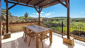 Une terrasse extérieure couverte, avec une table et des chaises, donnant sur un paysage verdoyant et bordée d’un mur en pierre.