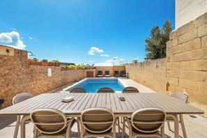 A poolside area with a table and chairs, a stone wall, and a clear blue sky.