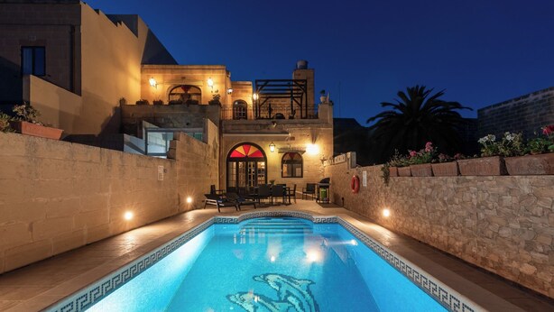 Une piscine décorée de motifs de dauphins, un mur en pierre et un bâtiment doté d’un balcon et d’éclairages.