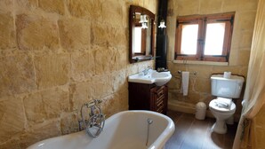 A bathroom with a stone wall, a bathtub, a sink, a mirror, a window, and a toilet. - Villeleynah Holiday Home (Gozo)