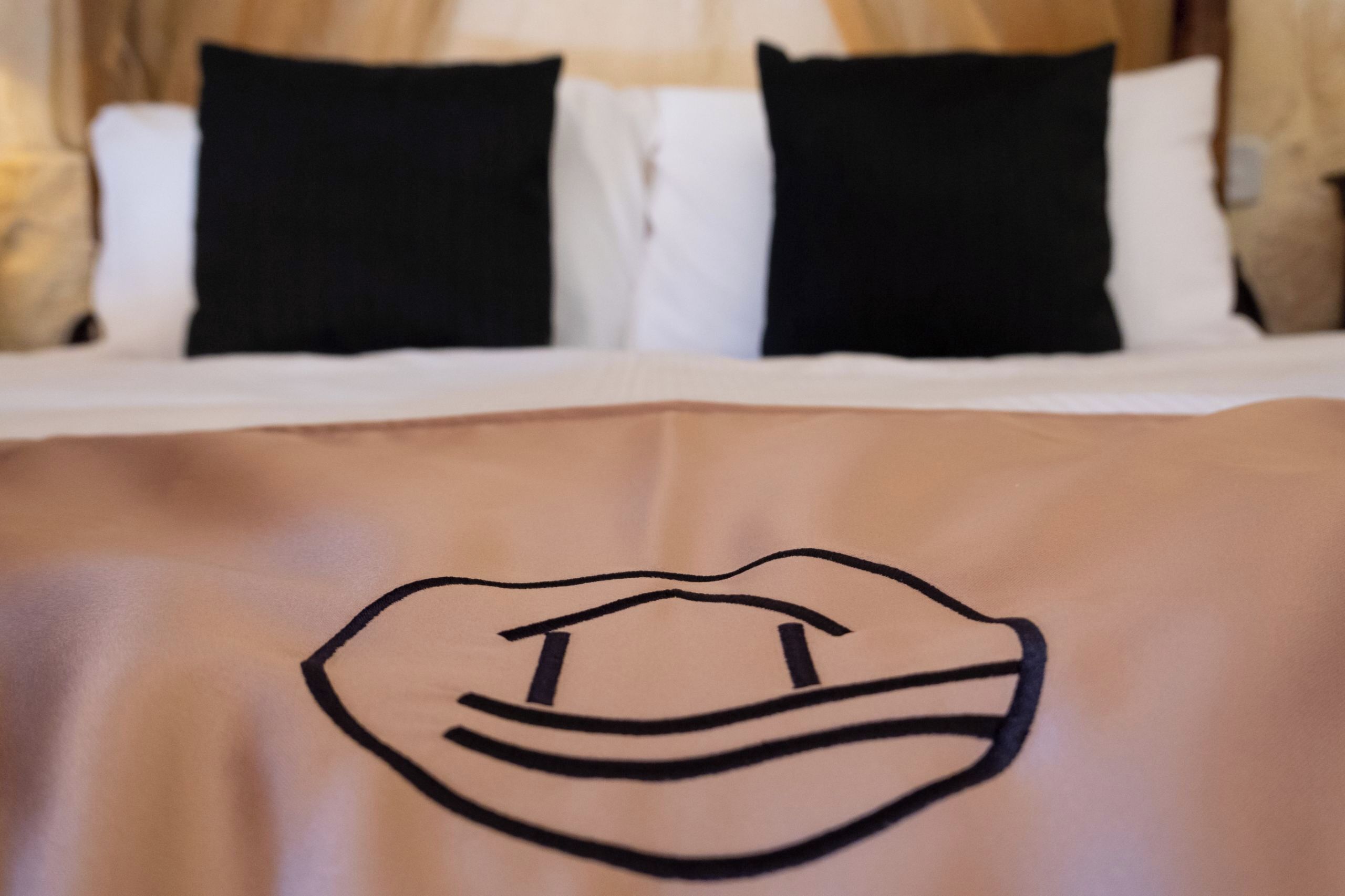 A bed with white and black pillows, a beige blanket with a black embroidered design, and a wooden headboard.