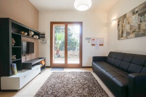 A modern living room with a black leather sofa, a flat-screen TV on a built-in shelf, a large abstract painting, and a sliding glass door leading to a garden.
