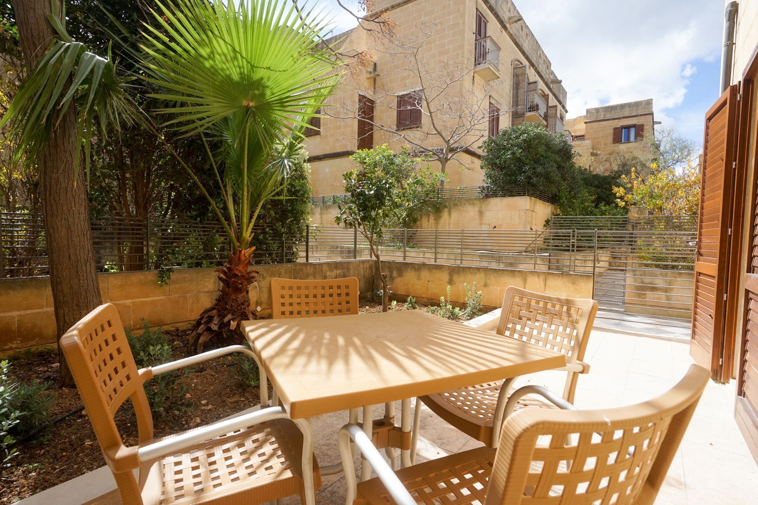 Eine Terrasse im Freien mit Tisch und Stühlen, umgeben von Pflanzen und einem Gebäude im Hintergrund.