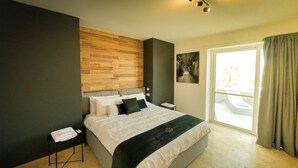 A modern bedroom with a large bed, a wooden accent wall, a nightstand, a bedside table, and a view of the outdoors through a sliding glass door. - Hygge Holiday Home (Ix-Xagħra)