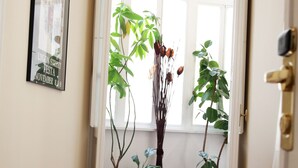Um corredor com piso ladrilhado, uma janela grande com molduras brancas e várias plantas em vasos.