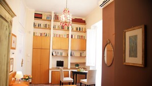 Quarto de hotel com cama, escrivaninha com cadeira, estante de livros, lustre e quadro na parede.