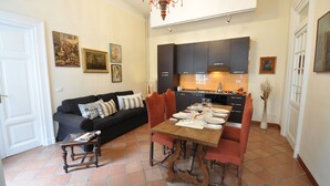 Uma área de refeições com uma mesa de madeira posta para quatro pessoas, um sofá preto com almofadas estampadas e uma cozinha ao fundo.