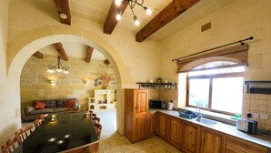 A spacious kitchen with a dining area, stone walls, wooden beams, and a large window. - Centre Island Holiday Home (Ir-Rabat Għawdex)