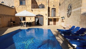 A swimming pool with lounge chairs, a table with chairs, and a covered seating area. - Centre Island Holiday Home (Ir-Rabat Għawdex)