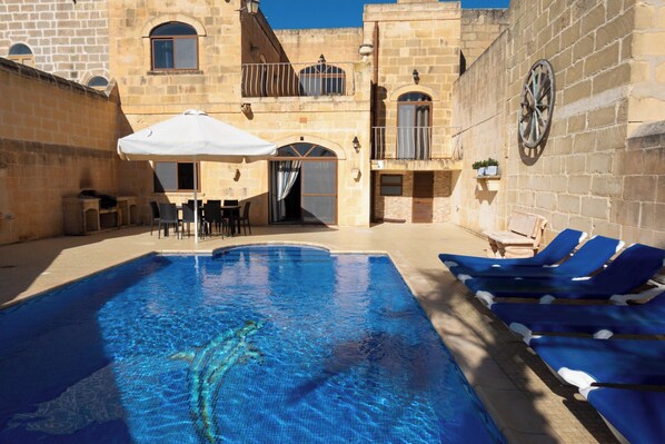 A swimming pool with lounge chairs, a table with chairs, and a covered seating area. - Centre Island Holiday Home (Ir-Rabat Għawdex)