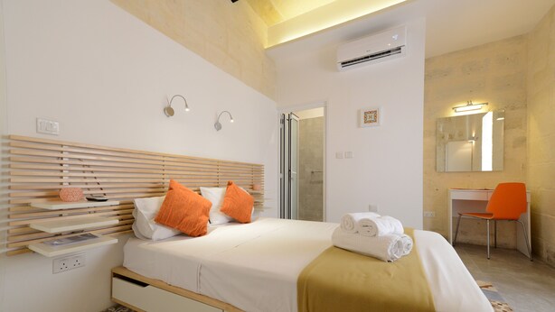 Ein modernes Hotelzimmer mit einem Holz-Kopfteil, einem Bett mit weißer Bettwäsche und orangefarbenen Kissen, einem Schreibtisch mit Stuhl sowie einem Badezimmer mit Spiegel und Dusche.