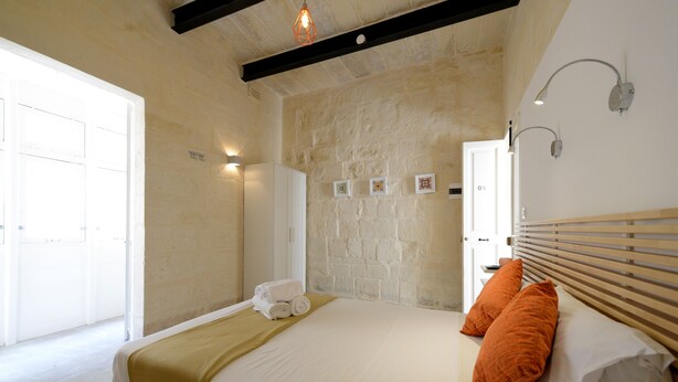 Un dormitorio con pared de piedra, cama con ropa de cama blanca y almohadas anaranjadas, cabecera de madera y puerta blanca.