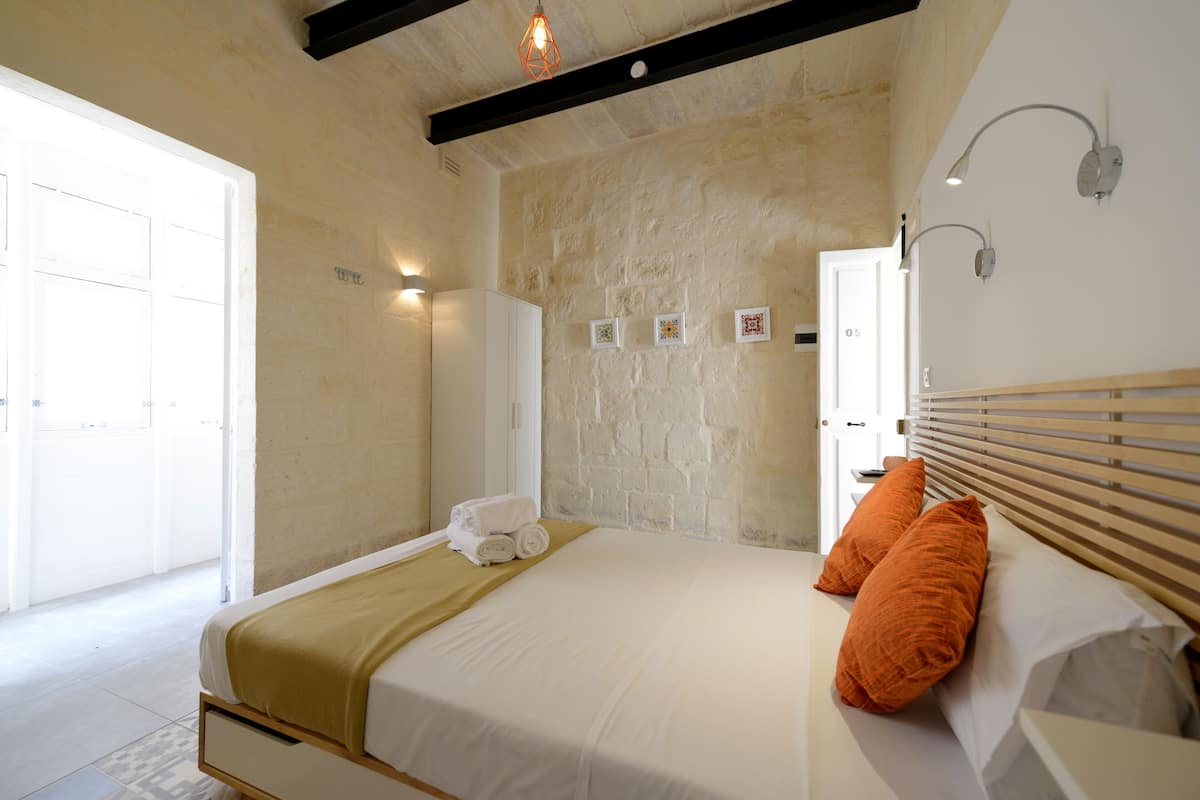 Un dormitorio con pared de piedra, cama con ropa de cama blanca y almohadas anaranjadas, cabecera de madera y puerta blanca.