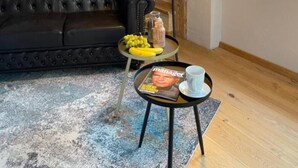 Een loft-achtige kamer met zichtbare houten balken, een leren bank, een kleine ronde tafel met een tijdschrift, een fruitschaal en een vloerlamp.