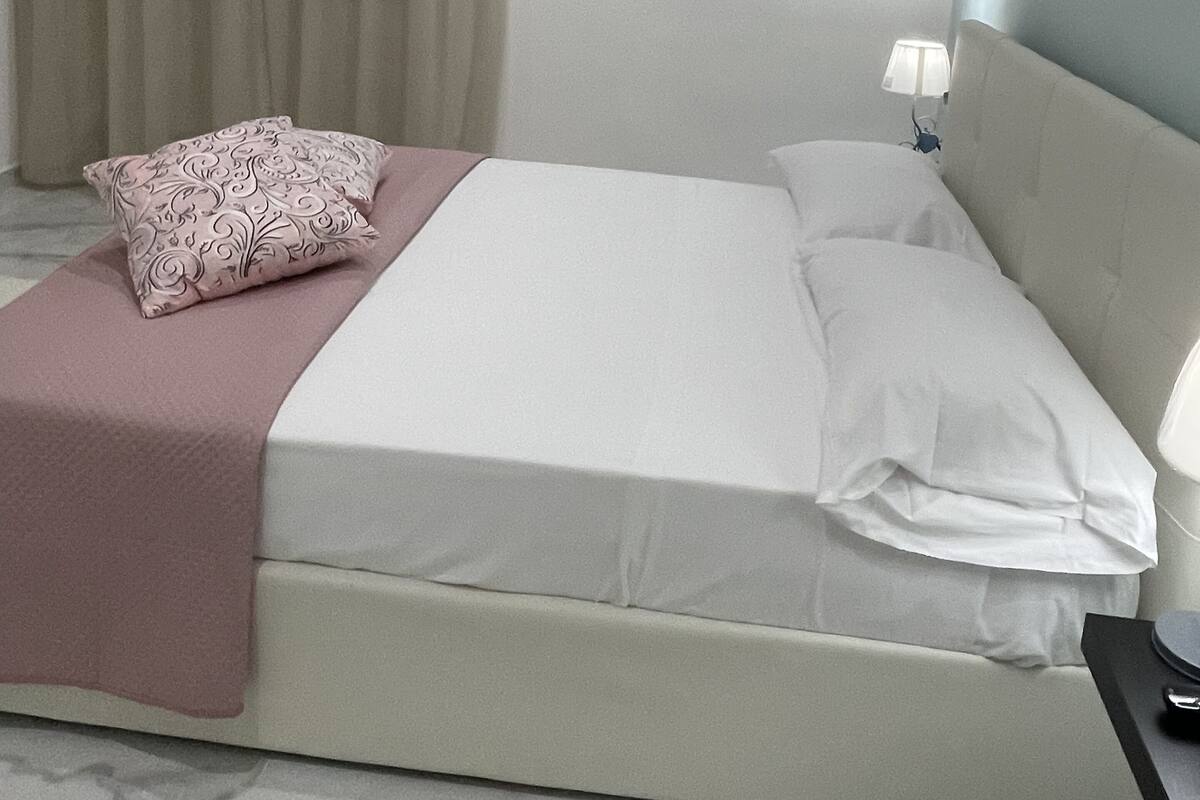 Ein ordentlich bezogenes Bett mit einem Kissen in Rosa und Weiß sowie einer weißen Decke und einer Nachttischlampe.