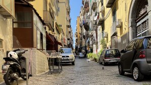 Una estrecha calle adoquinada flanqueada por edificios de varios pisos, coches aparcados y una moto.