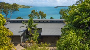 A building with a flat roof, surrounded by lush greenery and overlooking a calm sea with boats. - Shorelines 12 (Hamilton Island)