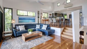 A modern living room with a blue sectional sofa, wooden coffee table, and a large painting of a beach. - Shorelines 12 (Hamilton Island)