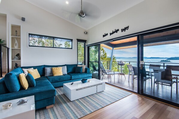 A modern living room with a teal sofa, a white coffee table, and a large sliding glass door leading to a balcony with a view of the water.