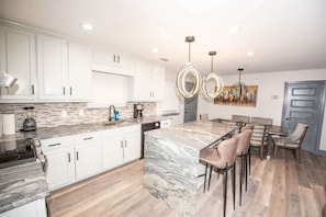 A modern kitchen with white cabinets, a marble island, and a dining area with wooden chairs.