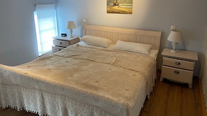 Une chambre avec un lit en bois, une literie blanche, deux tables de chevet avec des lampes, un tableau au mur et une fenêtre avec des stores.