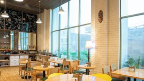A café with round wooden tables and yellow chairs, large windows, and a brick wall with a mural. - Bloom Hotel - Jammu (Jammu)