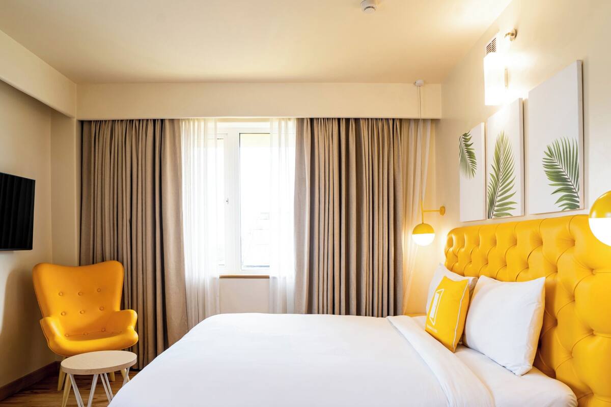 A hotel room with a large bed, a yellow armchair, a small round table, and a window with curtains.