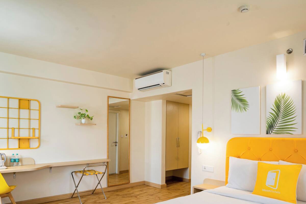 A modern hotel room with a wooden floor, a bed with a yellow headboard, a desk with a chair, a mirror, and a potted plant.