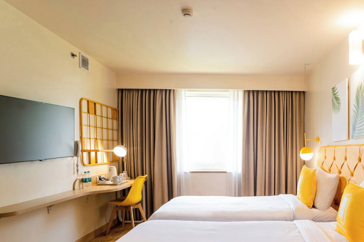 A hotel room with two beds, a desk with a chair, a flat-screen TV, a window with curtains, and a ceiling light.