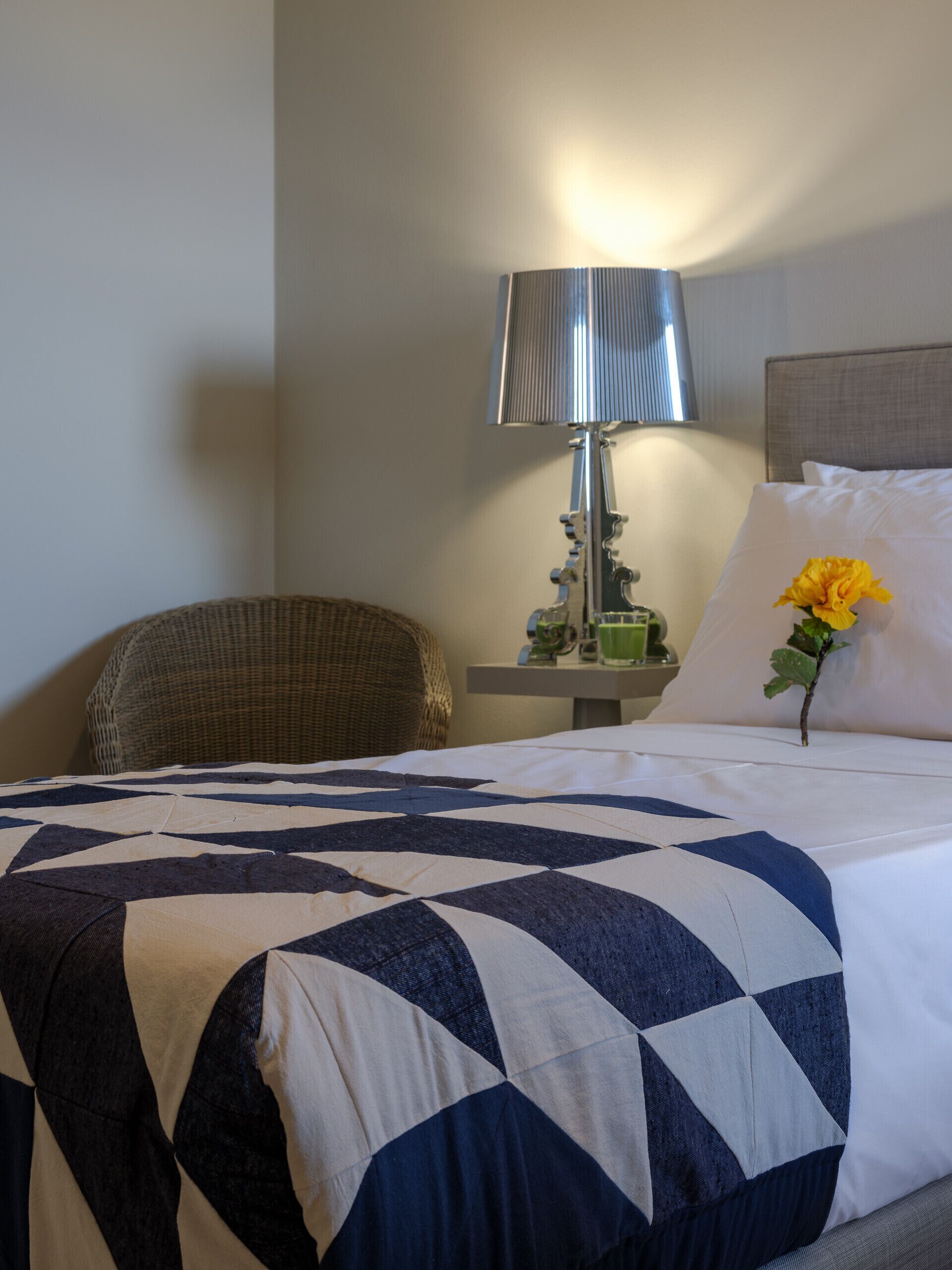 A neatly made bed with a geometric-patterned bedspread, a bedside lamp, a small table with a green vase, and a chair.
