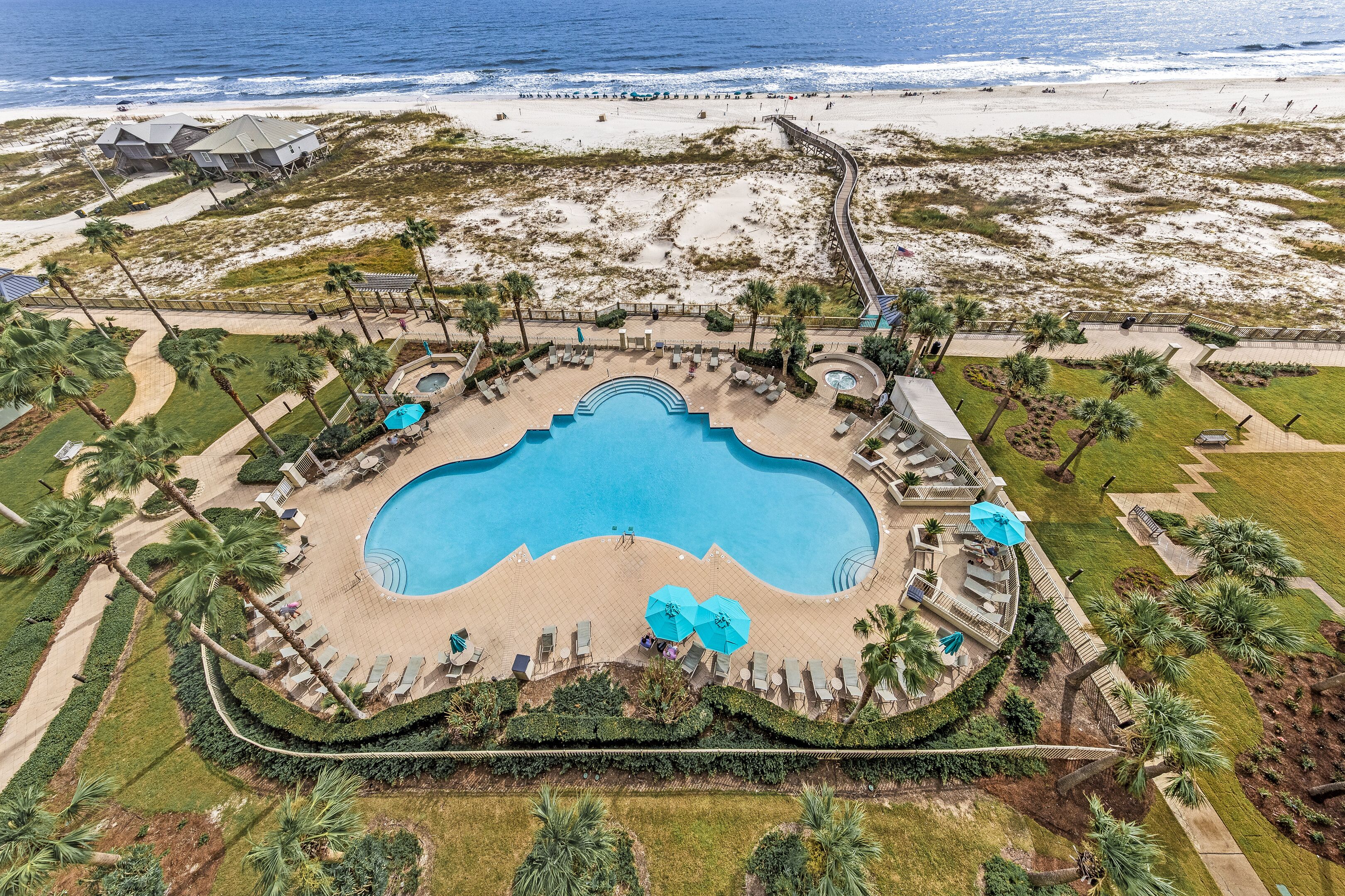 An aerial view of a resort with a central swimming pool, palm trees, and a beach in the background.