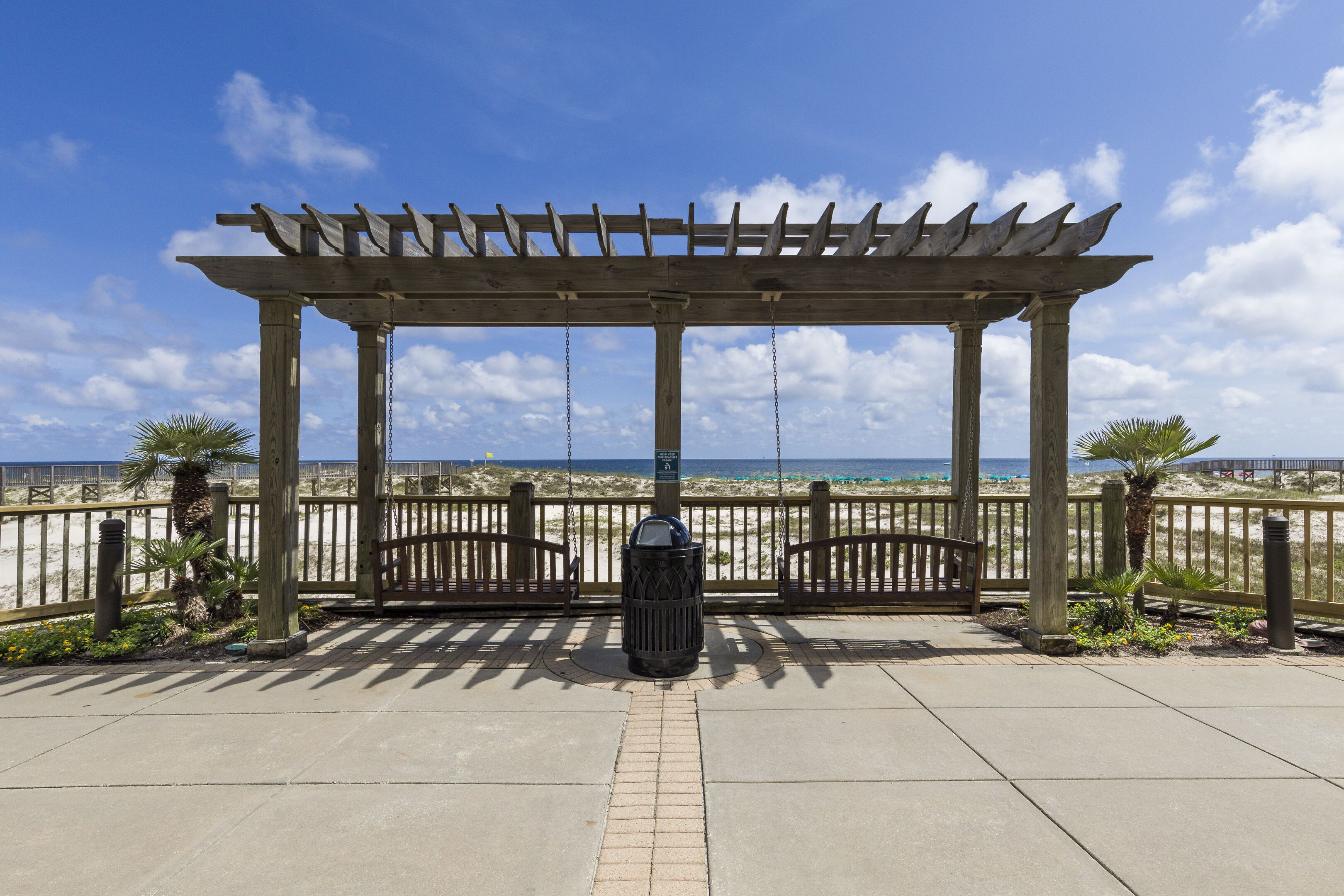 A wooden pergola with a view of the beach, a trash can, and a paved walkway.