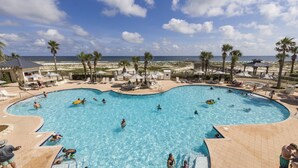 En stor, buet swimmingpool med folk, der svømmer og leger, omgivet af palmer og udsigt til stranden.