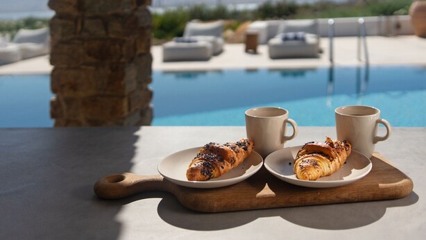Een tafel met twee croissants op borden, twee witte bekers en een houten plank. Achtergrond toont een zwembad en ligstoelen.
