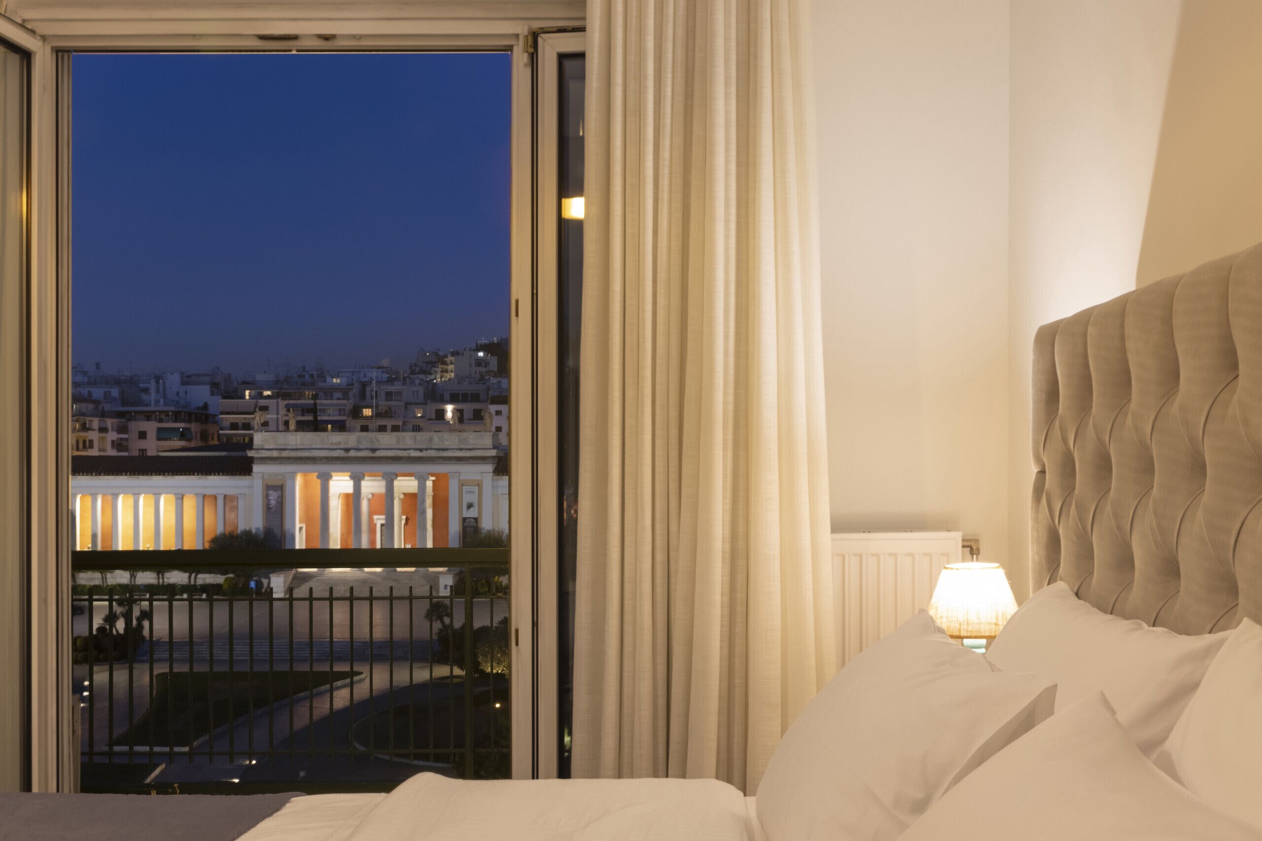A hotel room with a large window, a view of a cityscape, a bed with white linens, and a nightstand with a lamp.