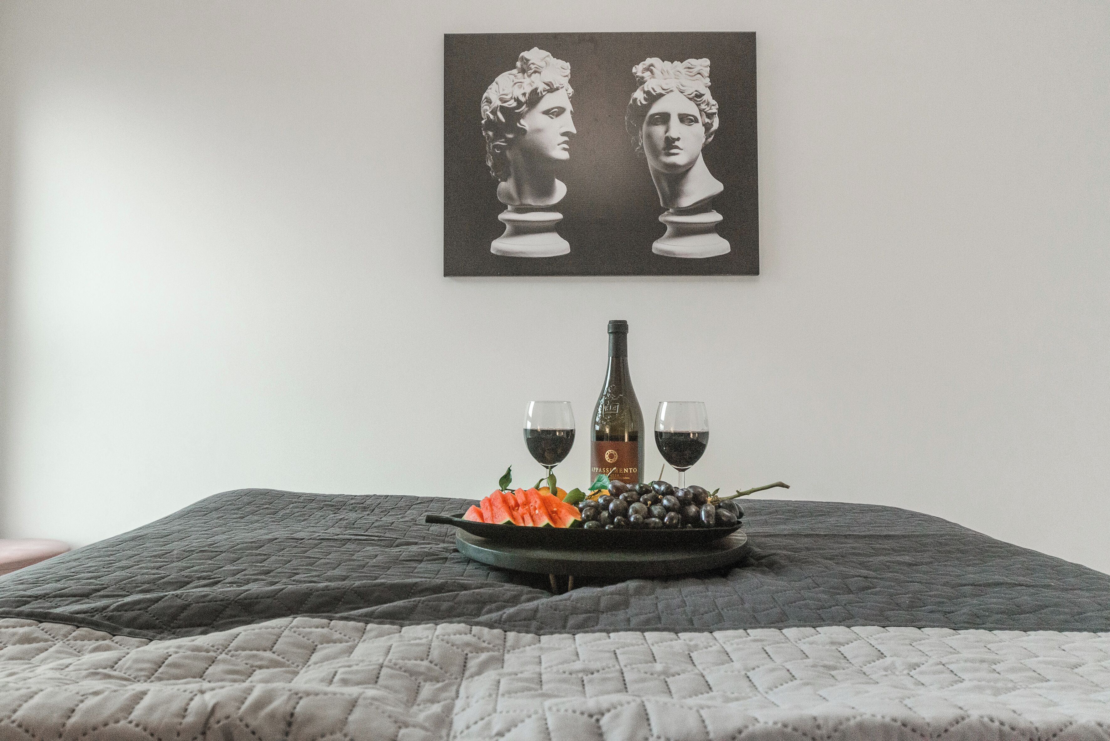 A bed with a quilted blanket, a black tray with grapes, a bottle, and two glasses of red wine on a table.