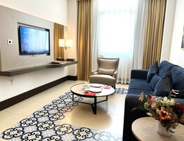 Una habitación de hotel moderna con una televisión de pantalla plana montada en la pared, un sofá, una silla, una mesa de centro y un jarrón decorativo con flores.