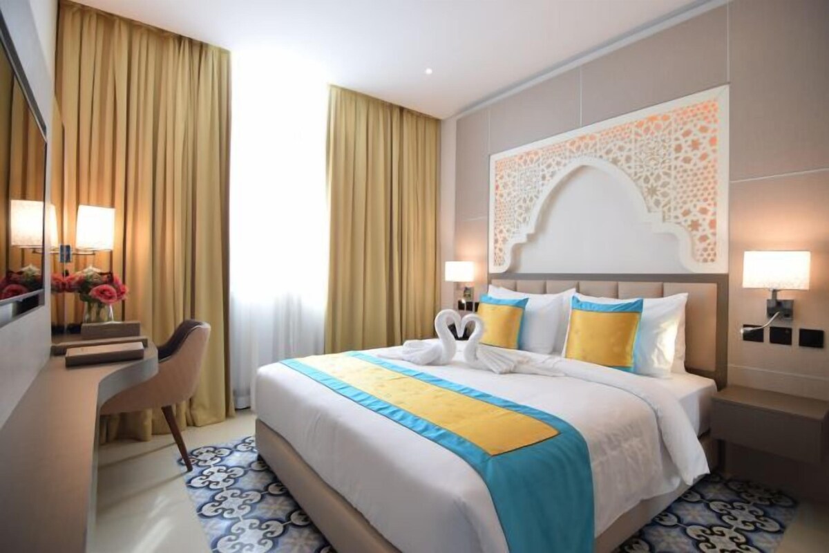 Une chambre d’hôtel avec un grand lit, deux lampes de chevet, un bureau avec une chaise et une grande fenêtre avec des rideaux.
