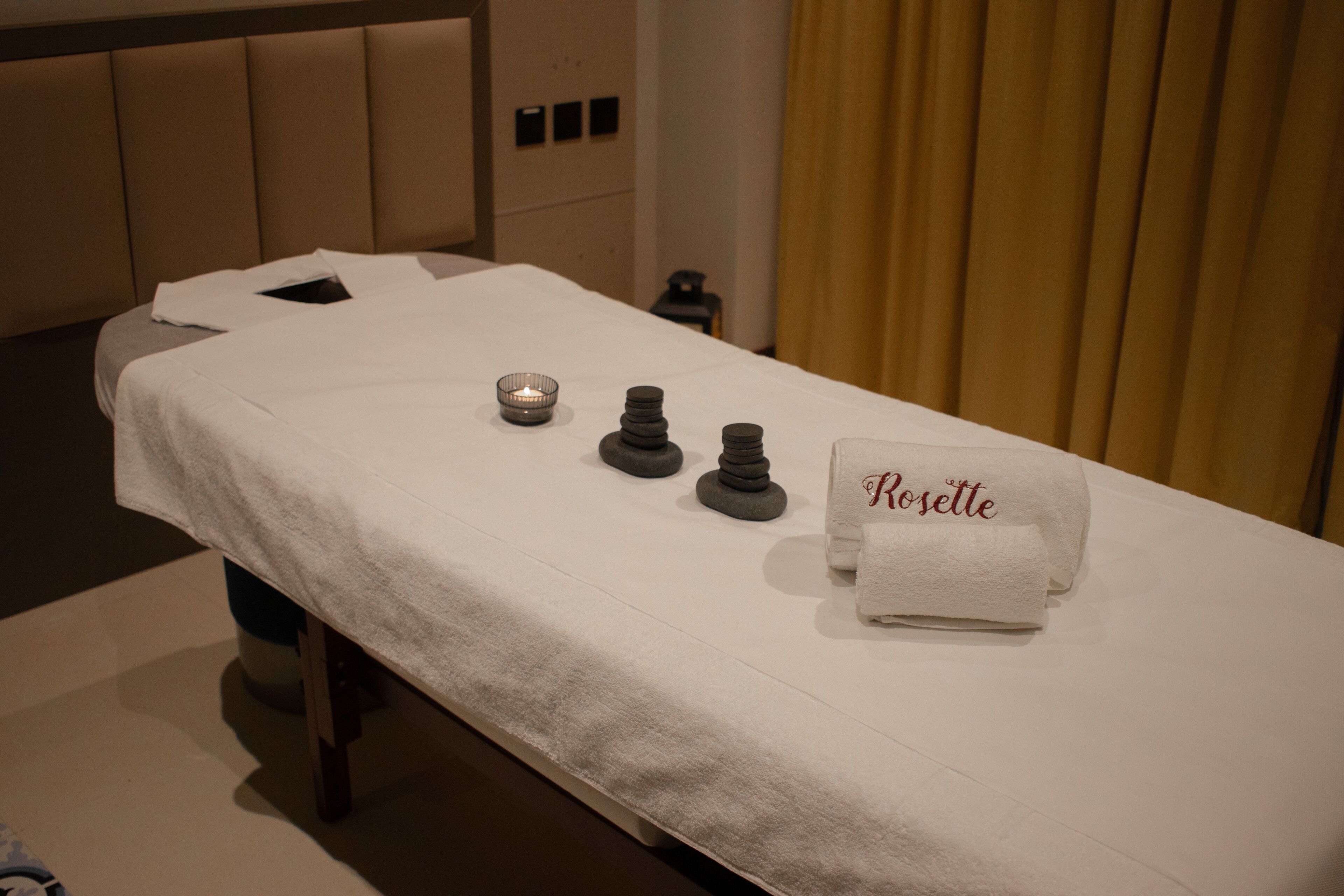A massage table with a white towel labeled "Rosette," a small candle, and a stack of stones.