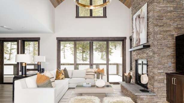 A spacious living room with a white sofa, fluffy ottomans, a fireplace, and large windows offering a view of trees. - Ski-In/Ski-Out | Hot Tub | Game Room (Big Sky)