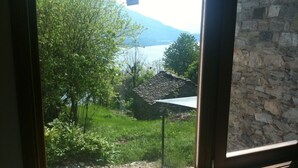 A view from a window with a stone wall and a radiator, overlooking a grassy area with trees and a building with a tiled roof. - La Campanelle (Montemezzo)